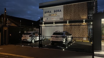 Un estacionamiento cerrado con dos coches dentro, un letrero sobre la entrada que dice "BORA BORA RESIDENCE", y un edificio con fachada de ladrillo.