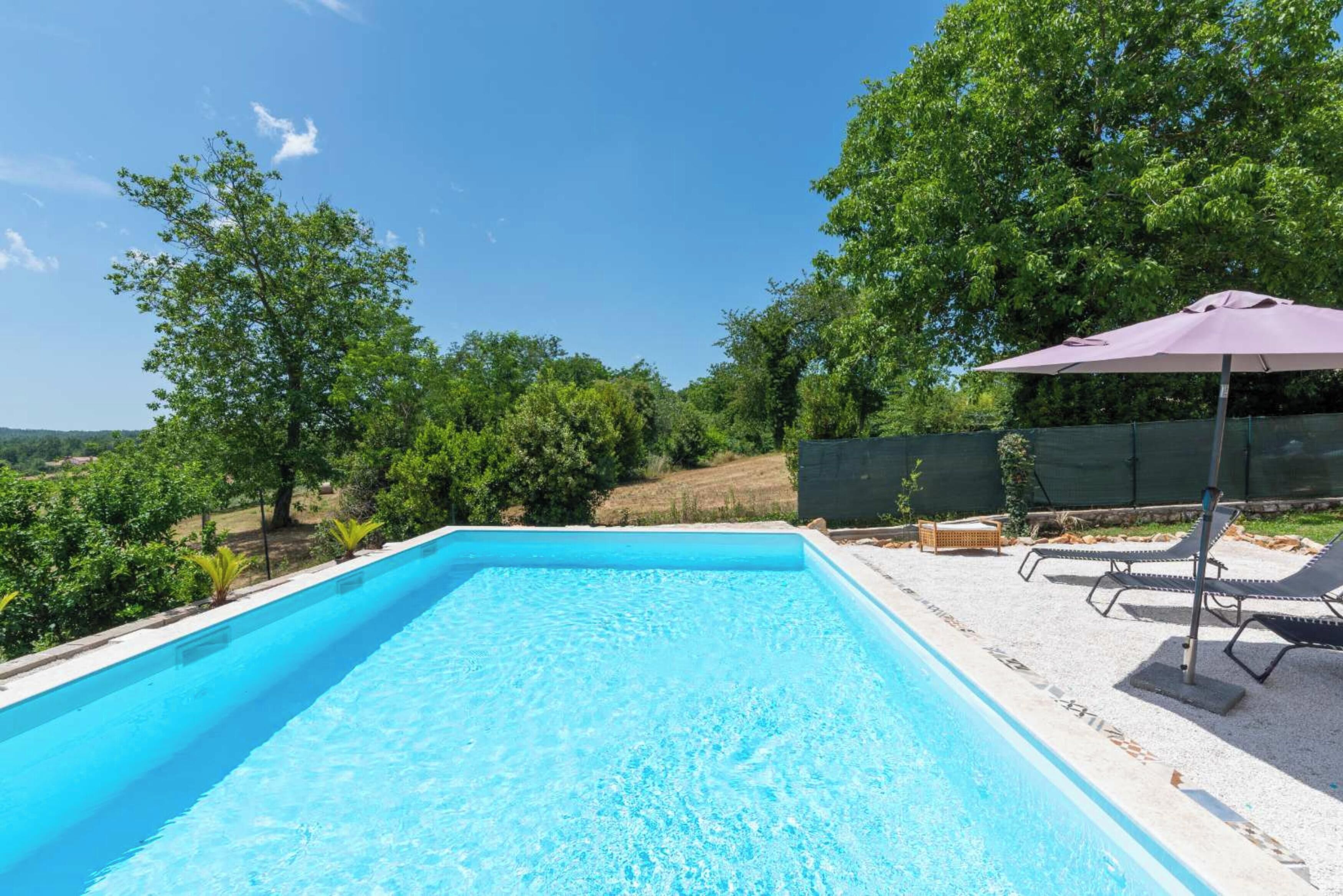 Et rektangulært pool med krystalklart blåt vand, omgivet af et grusareal med liggestole og en lilla parasol.