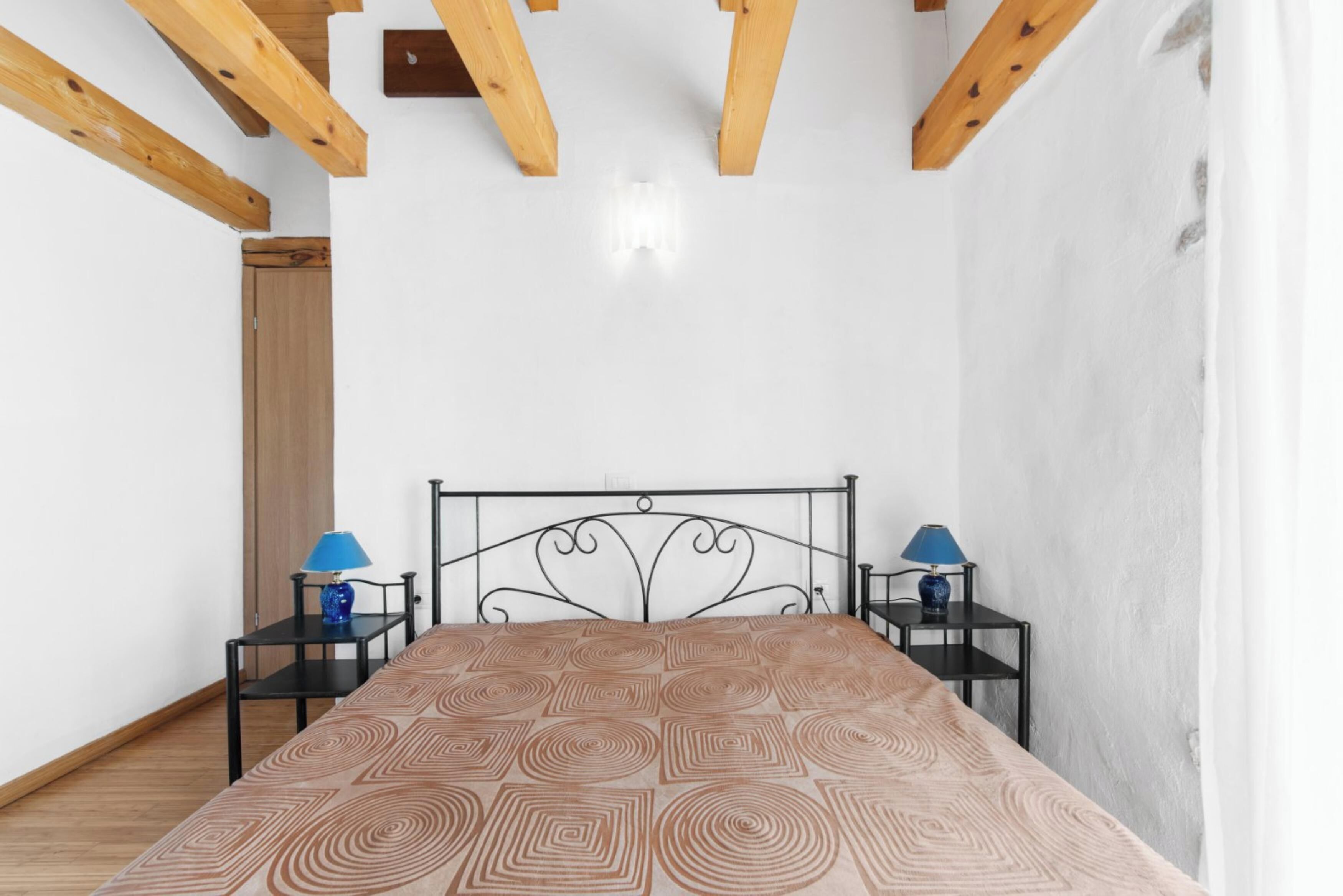 A bedroom with a bed, two bedside tables with lamps, wooden beams on the ceiling, and a white wall.