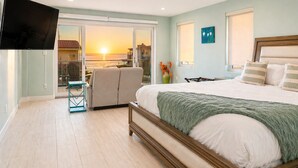 A bedroom with a bed, a TV, a balcony with a view of the sunset, and a chair. - 25% Off Aug 27-30 Walk 2 Beach, Amazing Ocean View (Oxnard)