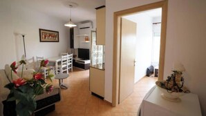 Una sala de estar con un sofá, una mesa de centro y un televisor. Se observa un comedor con una mesa y sillas. Se puede ver un área de cocina con un refrigerador y un microondas.
