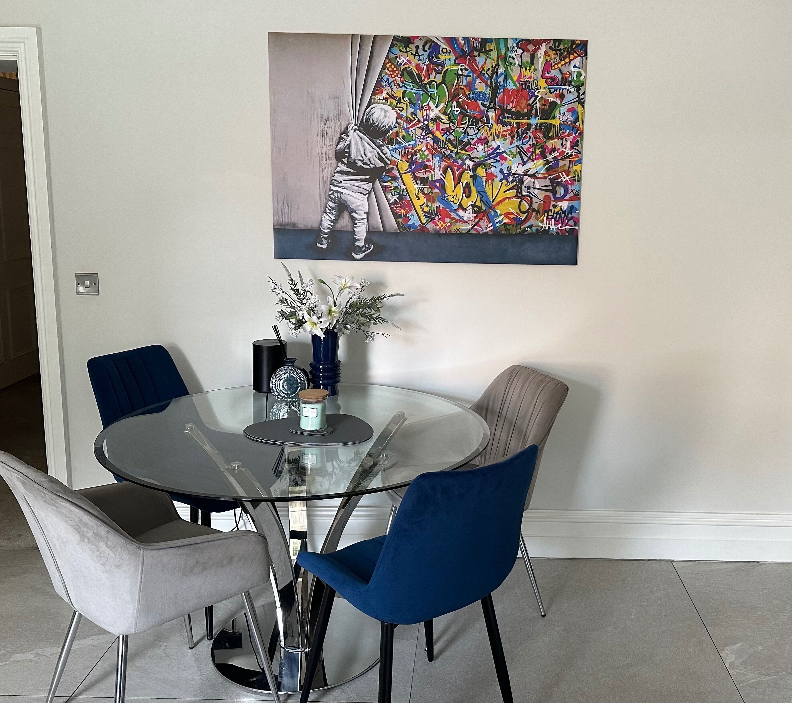 Un comedor con una mesa de vidrio rodeada de sillas, un jarrón con flores y un cuadro abstracto y colorido en la pared.