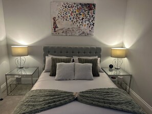 Une chambre à coucher avec une tête de lit capitonnée, un lit agrémenté de plusieurs coussins, deux tables de chevet avec des lampes et une œuvre d’art accrochée au mur.
