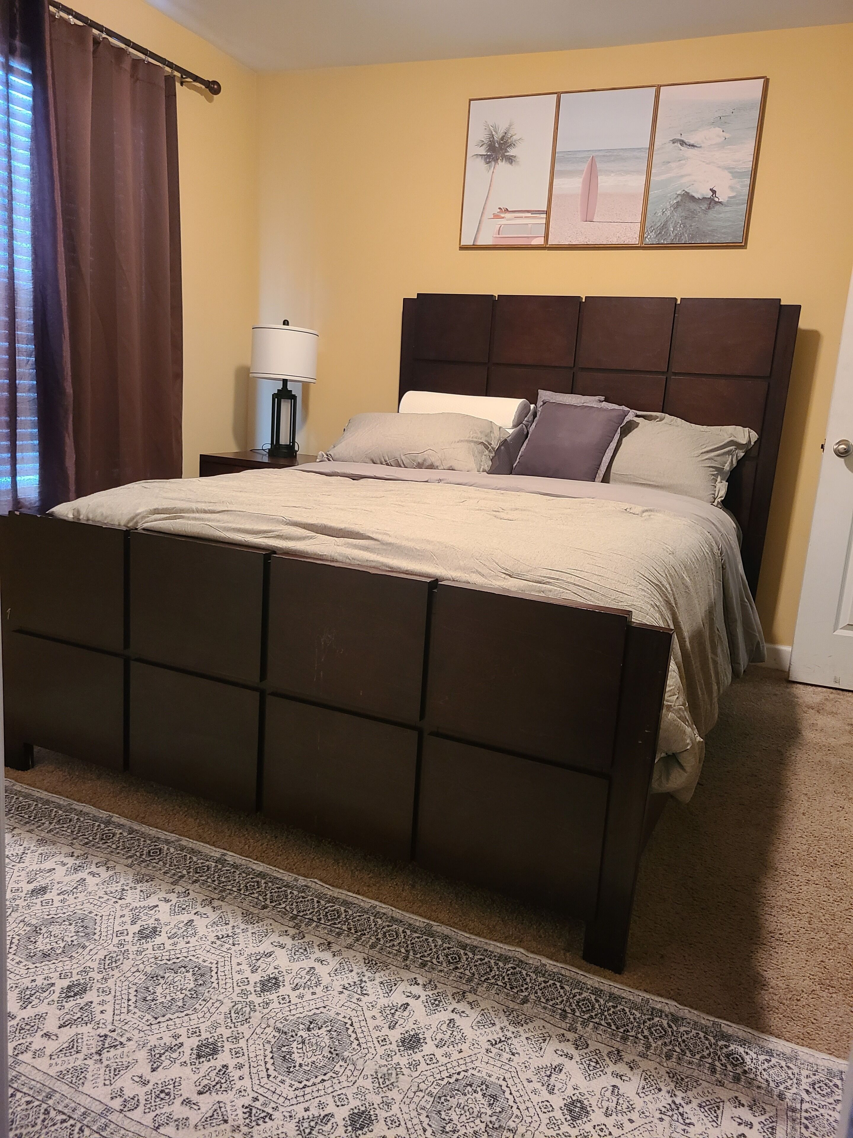 A bedroom with a bed, a nightstand, a lamp, a patterned rug, and a wall with three framed pictures.