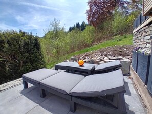 A patio with cushioned seating and a round table, surrounded by a stone wall and greenery. - Modern apartment in the glass town of Lauscha with terrace, beautiful nature & mountain views (Lauscha)