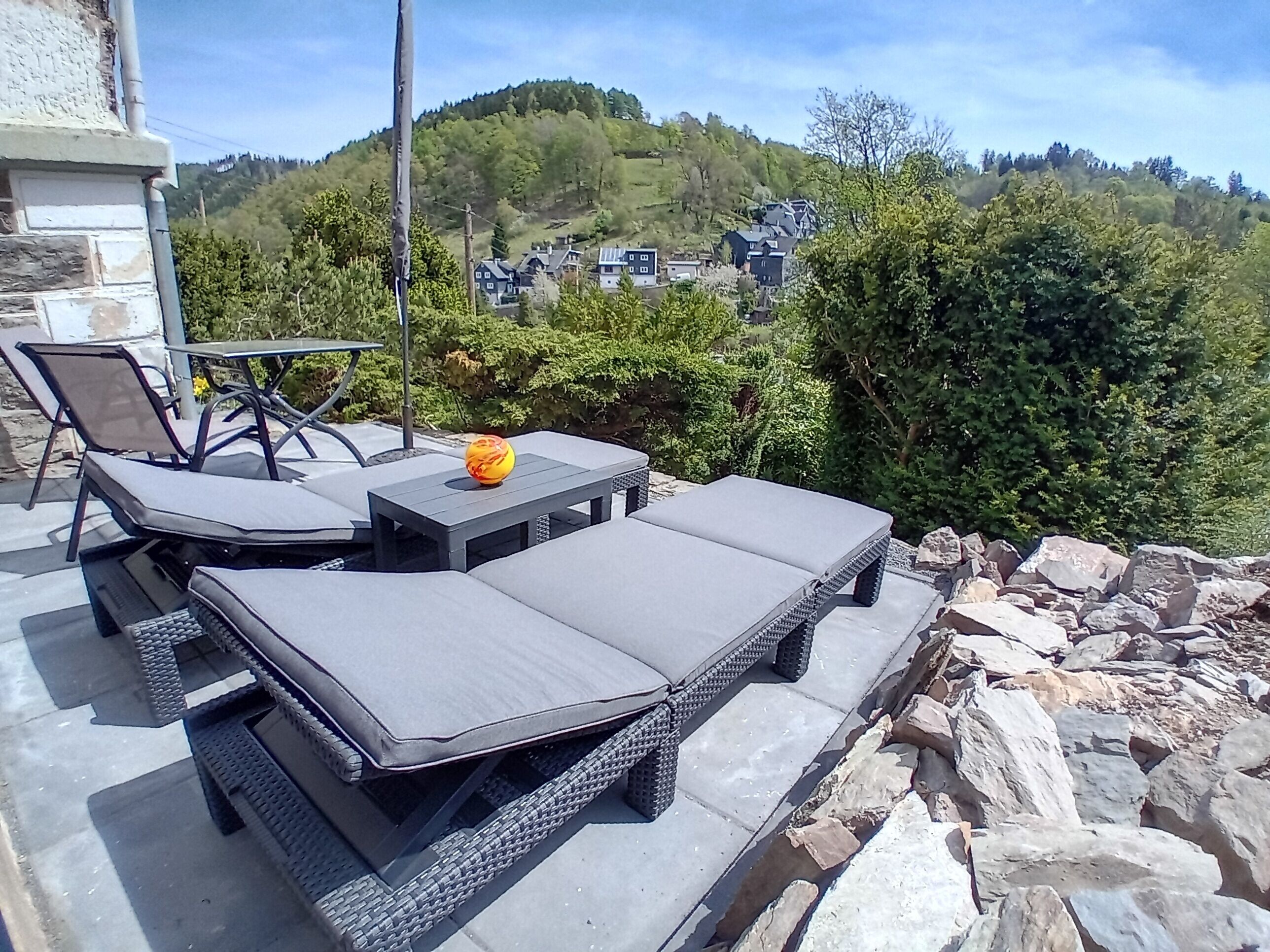 Ein Außenbereich mit Lounge-Möbeln und einem Tisch, umgeben von Grünpflanzen und Blick auf Häuser in der Ferne.