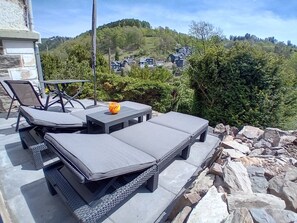An outdoor seating area with lounge chairs and a table, surrounded by greenery and a view of houses in the distance.