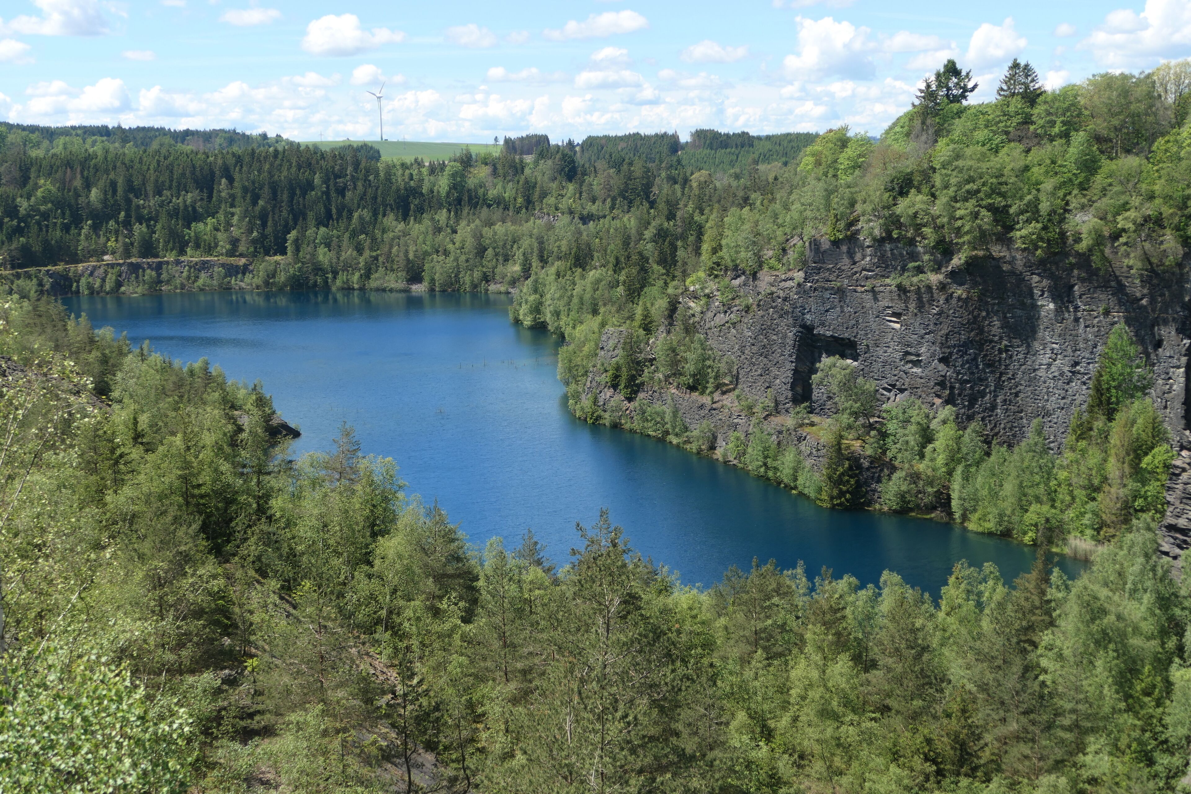 Un paisaje natural con un lago rodeado de bosque denso y un risco con la entrada de una cueva.