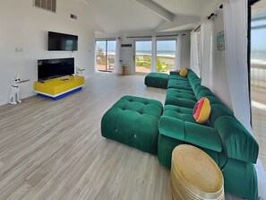 A modern living room with a large flat-screen TV, a yellow and blue entertainment unit, a green sectional sofa, and a wicker ottoman.