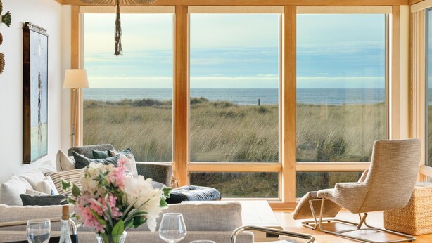 Habitación con un ventanal grande con vista a una playa, un sofá, una silla y una mesa de comedor con vasos y una botella.