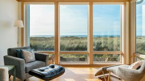 Amplia habitación con ventanas grandes que ofrecen vista a la playa y las dunas. Hay dos sillones y una mesita con una lámpara.