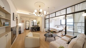 A modern living room with a sofa, a round coffee table, and a dining area with a table and chairs. There is a large window with curtains, a bed in the background, and a chandelier. - YIDU (Nanjing West Road Store) (Shanghai)