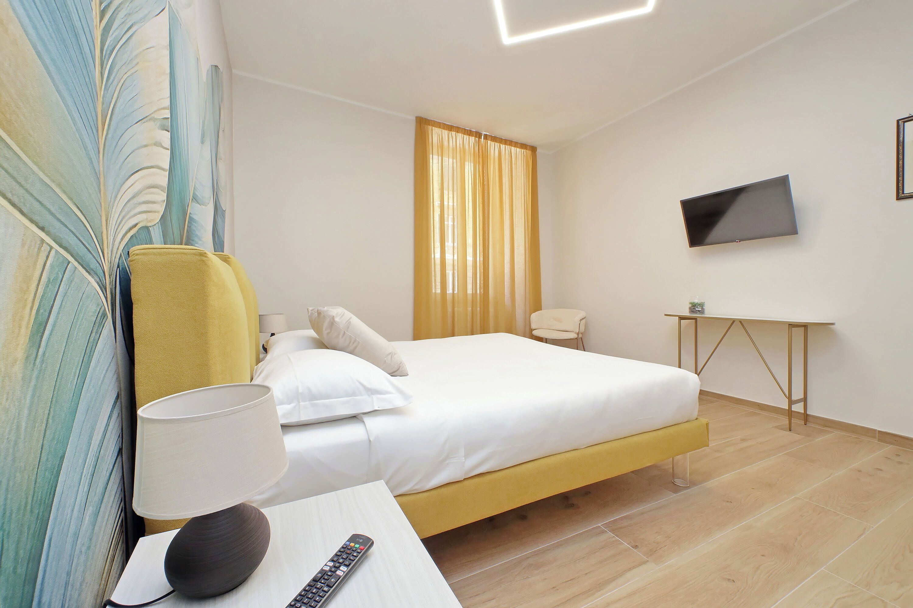 A hotel room with a bed, a TV mounted on the wall, a desk with a lamp, and a chair.