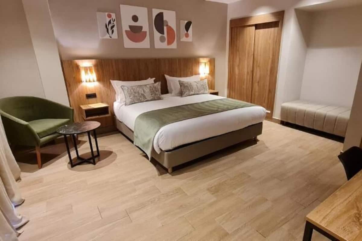 A modern bedroom with a large bed, a green armchair, a small table, and a wooden wardrobe.