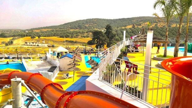Zona de parque acuático con un tobogán grande, piscina y vistas a un paisaje montañoso.
