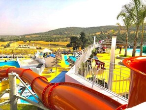 Ein Wasserparkbereich mit einer großen Rutsche, einem Schwimmbecken und Blick auf eine hügelige Landschaft.
