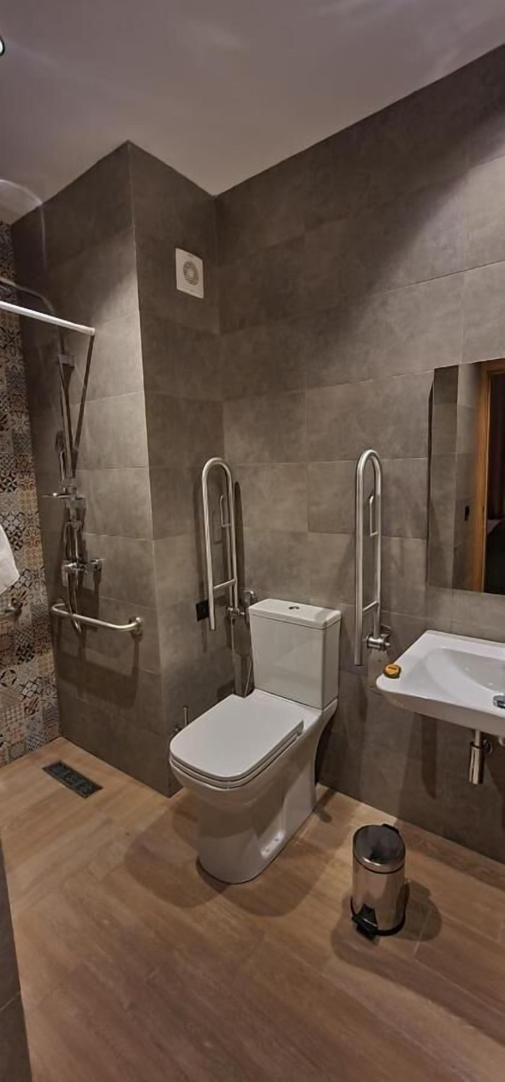 Un baño moderno con inodoro, bidé y lavamanos. Cuenta con una zona de ducha con puerta de cristal y barra de apoyo. Se observa un cubo de basura en el suelo.