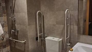 Un baño moderno con inodoro, bidé y lavamanos. Cuenta con una zona de ducha con puerta de cristal y barra de apoyo. Se observa un cubo de basura en el suelo.