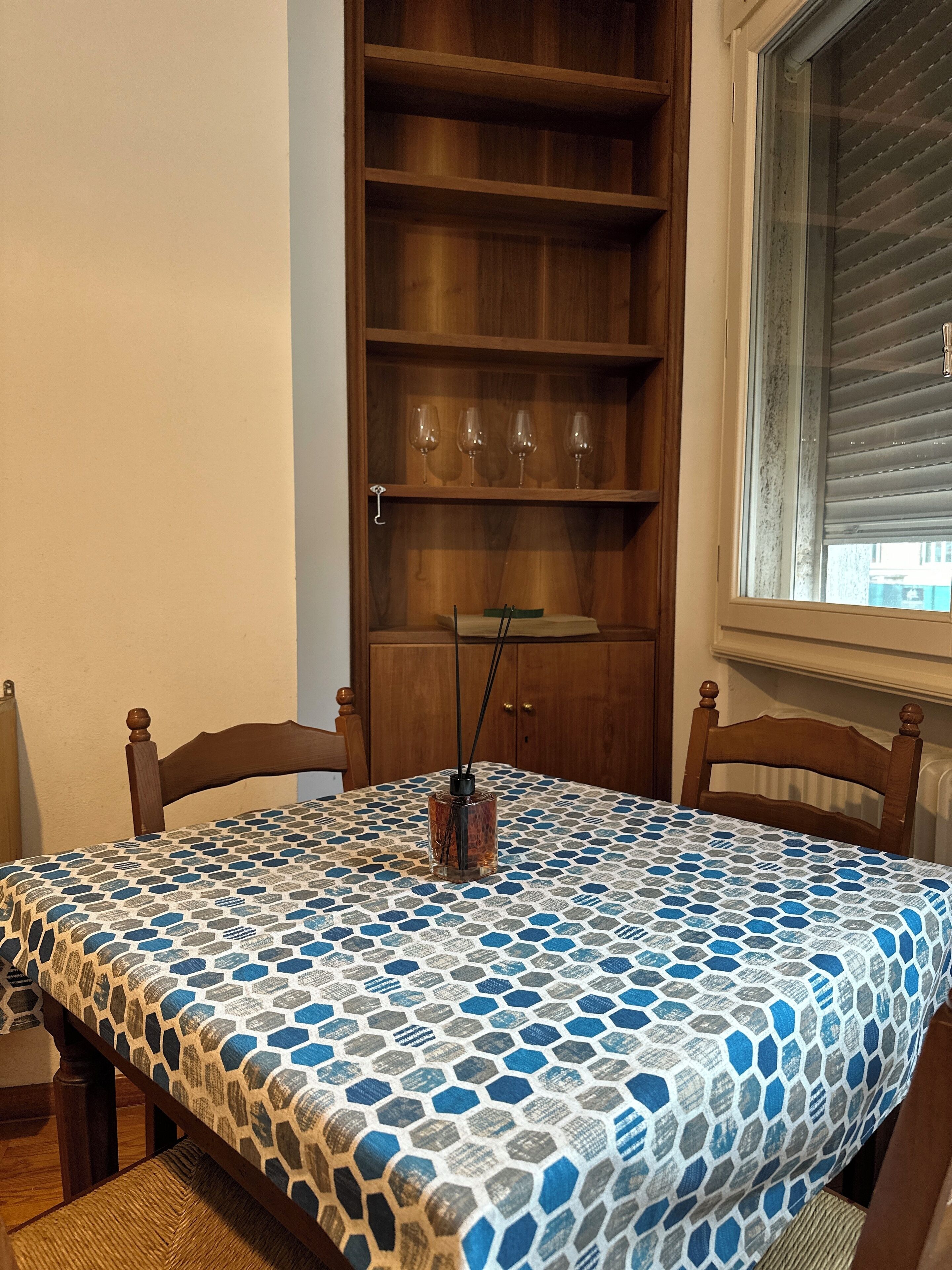 Een eettafel met een tafelkleed met patroon, houten stoelen, een kast met wijnglazen en een raam met jaloezieën.