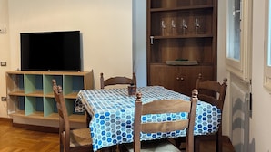 Un comedor con una mesa dispuesta para cuatro, sillas de madera, un televisor sobre un mueble bajo y un armario de madera con estantes.