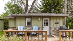 A small, single-story building with a front porch, two blue Adirondack chairs, a wooden table, and a barbecue grill. - Westlake Couple`s Cottage (Prince Edward)