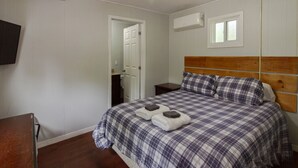 A bedroom with a bed, a wooden headboard, a flat-screen TV mounted on the wall, and an air conditioning unit. - Westlake Couple`s Cottage (Prince Edward)