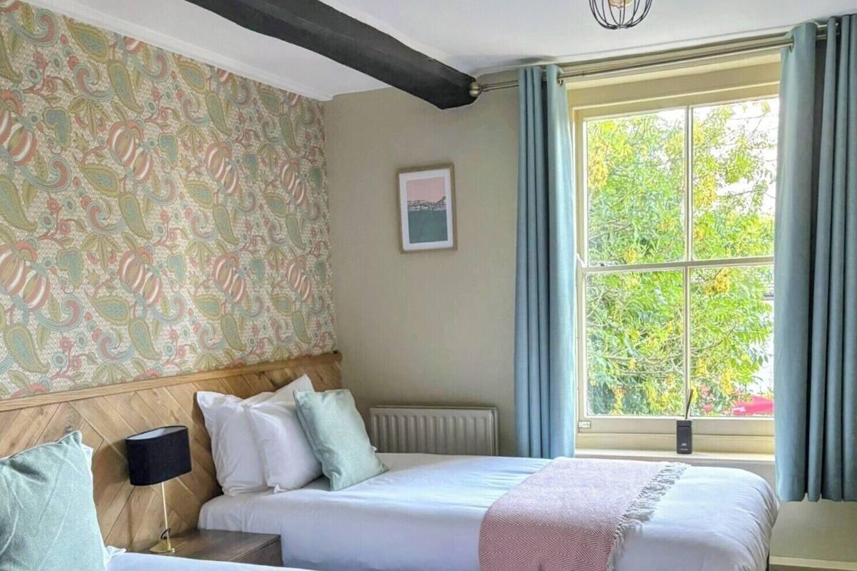 A hotel room with two beds, a large window with curtains, a patterned wallpaper, and a hanging light fixture.