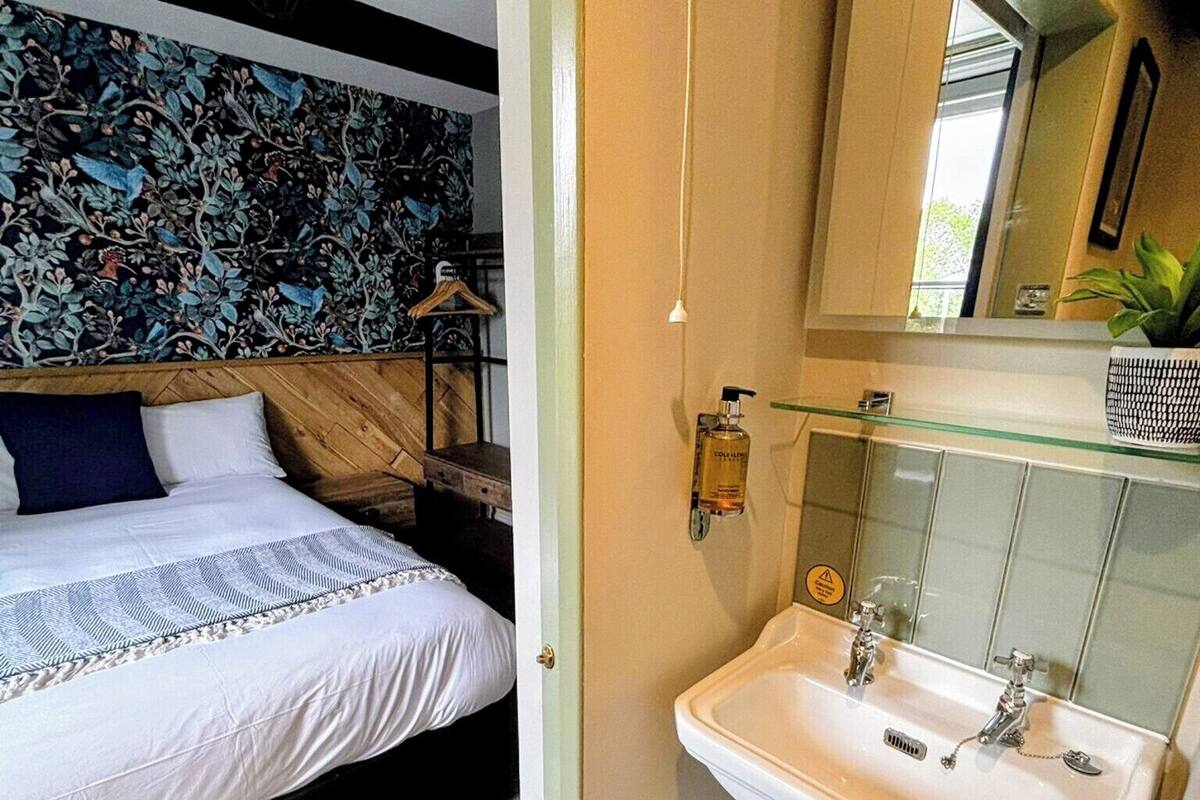 A bedroom with a bed, a nightstand, a wall-mounted lamp, and a bathroom with a sink and mirror.