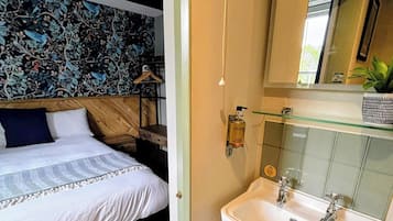 Une chambre à coucher comprenant un lit, une table de chevet et une lampe fixée au mur, ainsi qu’une salle de bain avec un lavabo et un miroir.