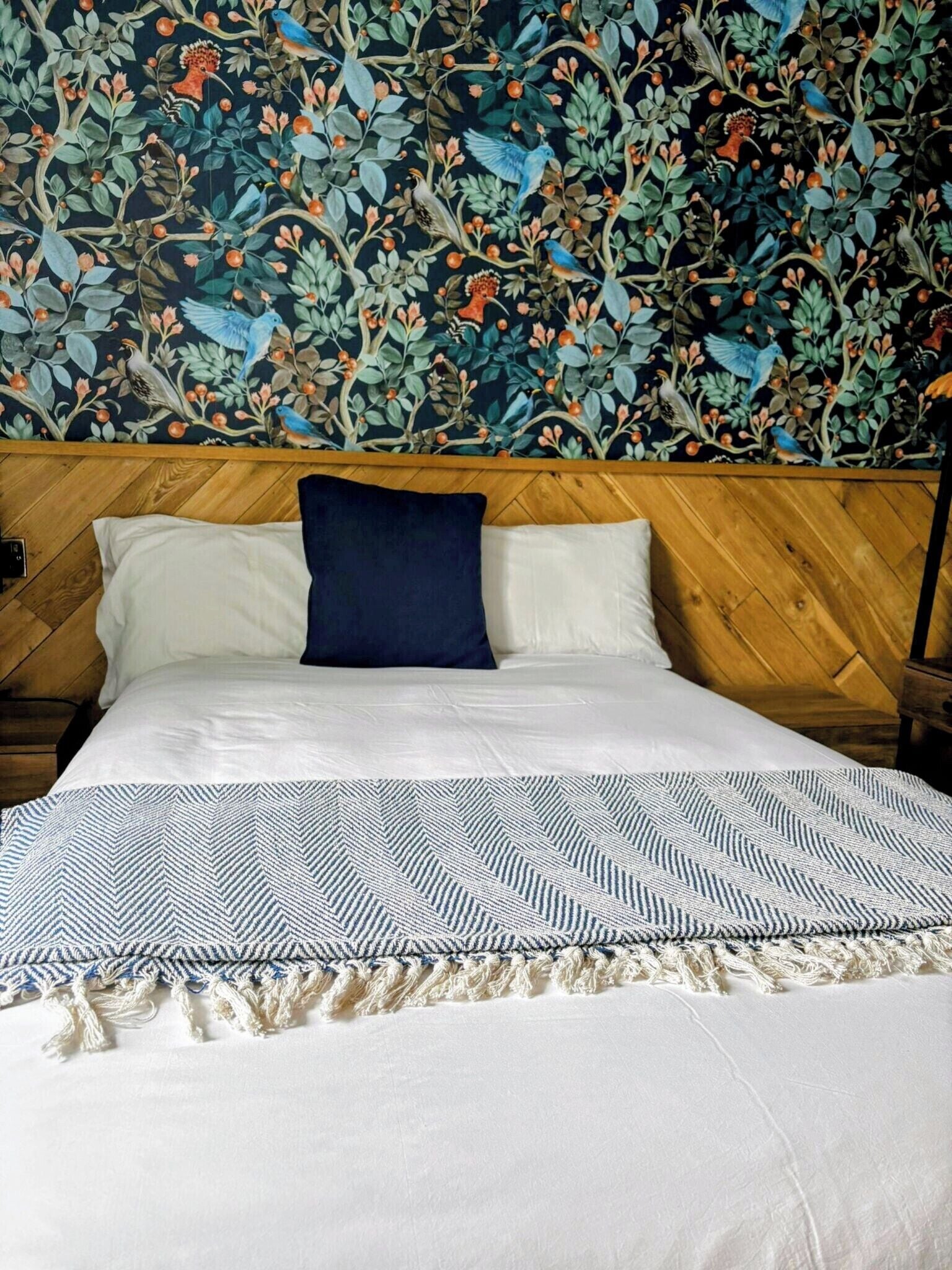 Una cama bien hecha con colcha y almohadas estampadas, cabecera de madera y pared con diseño de papel tapiz floral.