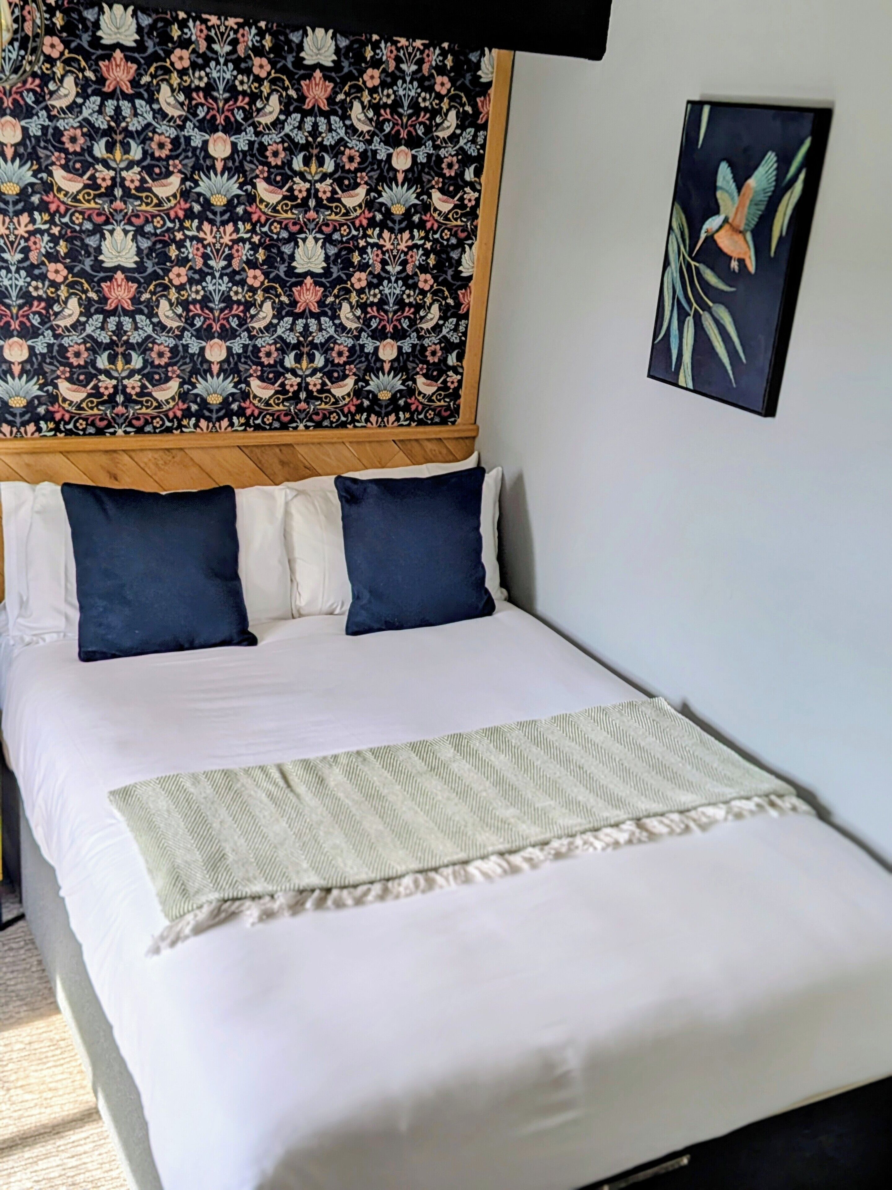 Una cama bien hecha con ropa de cama blanca y azul, un tapiz estampado encima y una obra enmarcada en la pared.