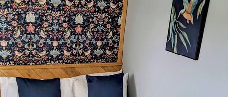 Una cama bien hecha con ropa de cama blanca y azul, un tapiz estampado encima y una obra enmarcada en la pared.