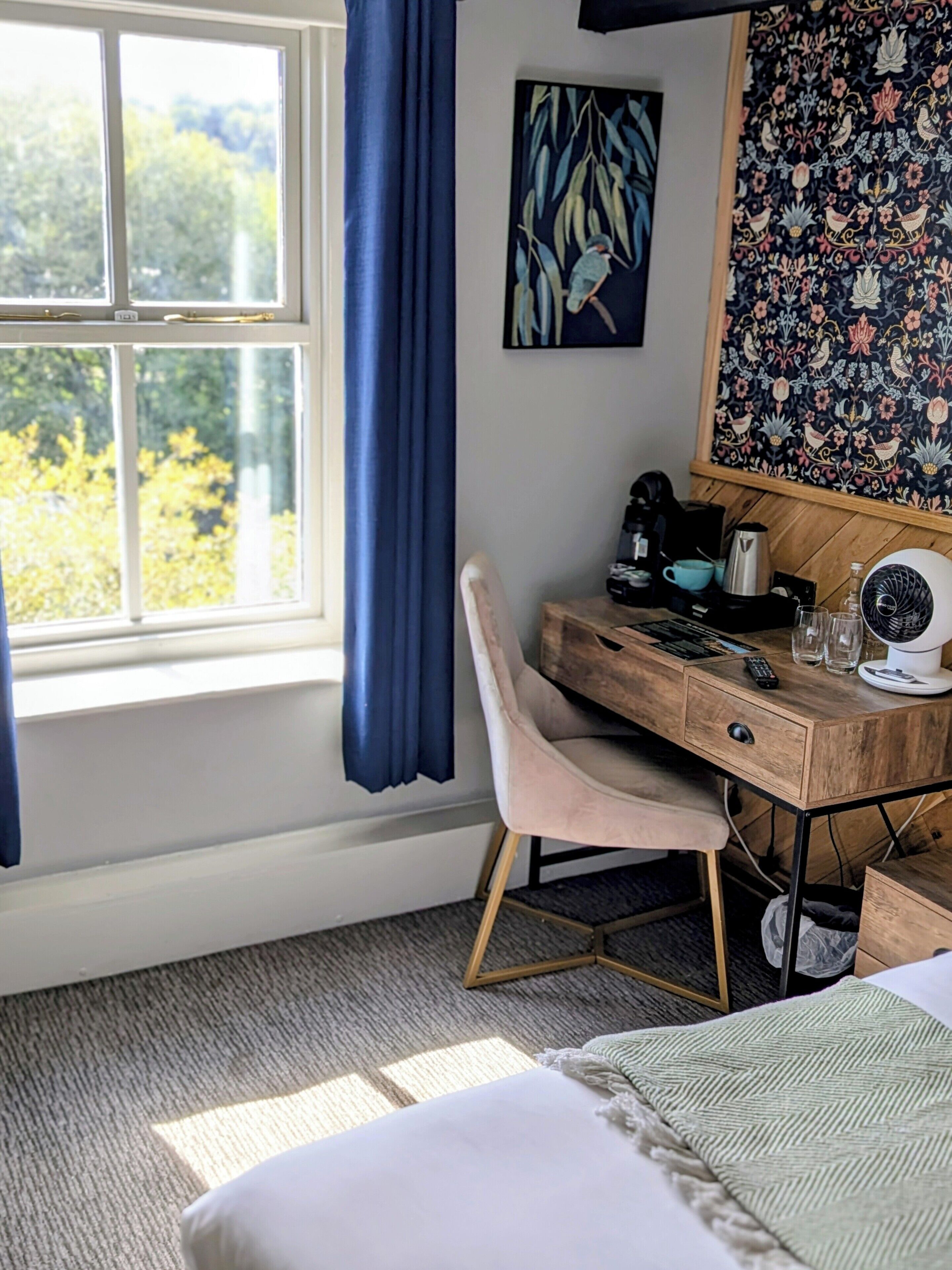 A bedroom with a bed, a desk with a chair, a window with curtains, a painting, and a patterned wall.