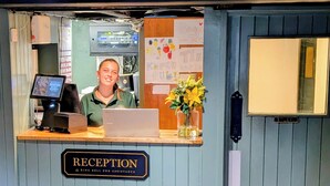 A reception desk with a sign reading "RECEPTION & RING BELL FOR ASSISTANCE," a computer, a vase with flowers, and a bulletin board with notes. - Old Mill Salisbury (Salisbury)