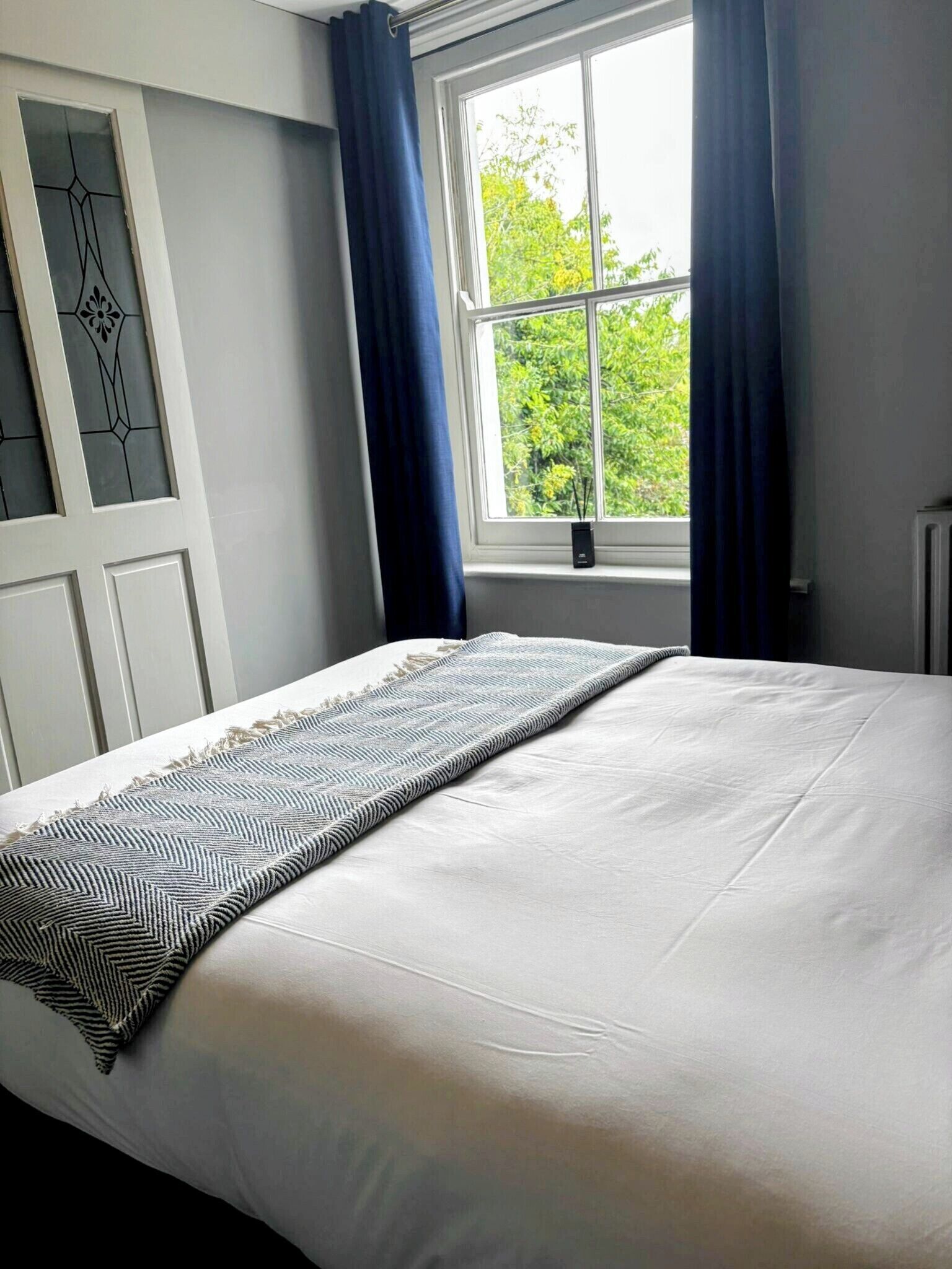 A neatly made bed with a patterned throw, a window with blue curtains, and a view of greenery outside.