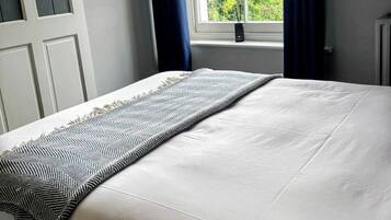 A neatly made bed with a patterned throw, a window with blue curtains, and a view of greenery outside.