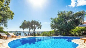 A swimming pool with a stone border, surrounded by lounge chairs and trees, with a view of the sea in the background. - Villa Jasmin Nissaki (Nisaki)