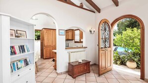 A well-lit room with a white bookshelf, wooden cabinets, a radiator, and a door leading to an outdoor area with plants and a view of the sky. - Villa Jasmin Nissaki (Nisaki)