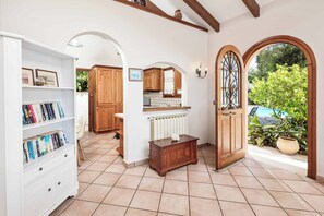 A well-lit room with a white bookshelf, wooden cabinets, a radiator, and a door leading to an outdoor area with plants and a view of the sky.