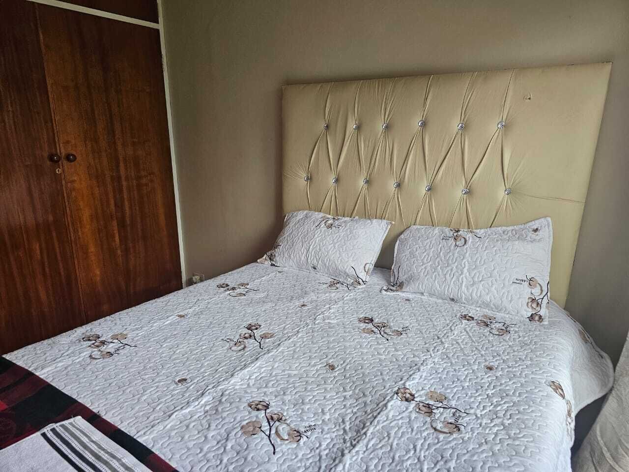 Un lit avec une tête de lit capitonnée, une literie blanche à motifs floraux et une armoire en bois sur le côté gauche.