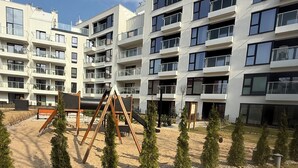 A modern apartment building with multiple balconies and a playground area with swings and small trees.