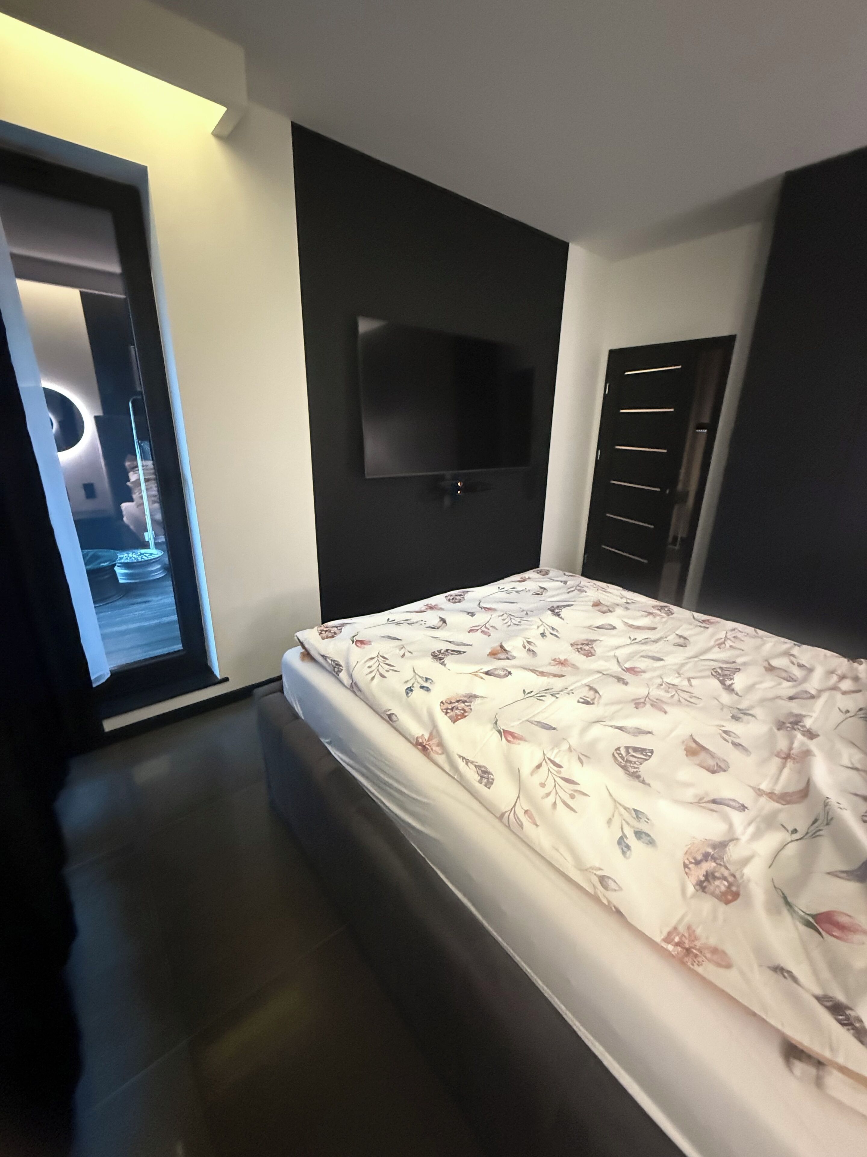 A bedroom with a bed, a flat-screen TV mounted on the wall, a door, and a window with a view of a circular light fixture.