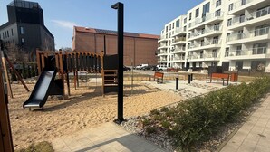 A playground with a slide and swings, a paved walkway, and a building with balconies in the background.