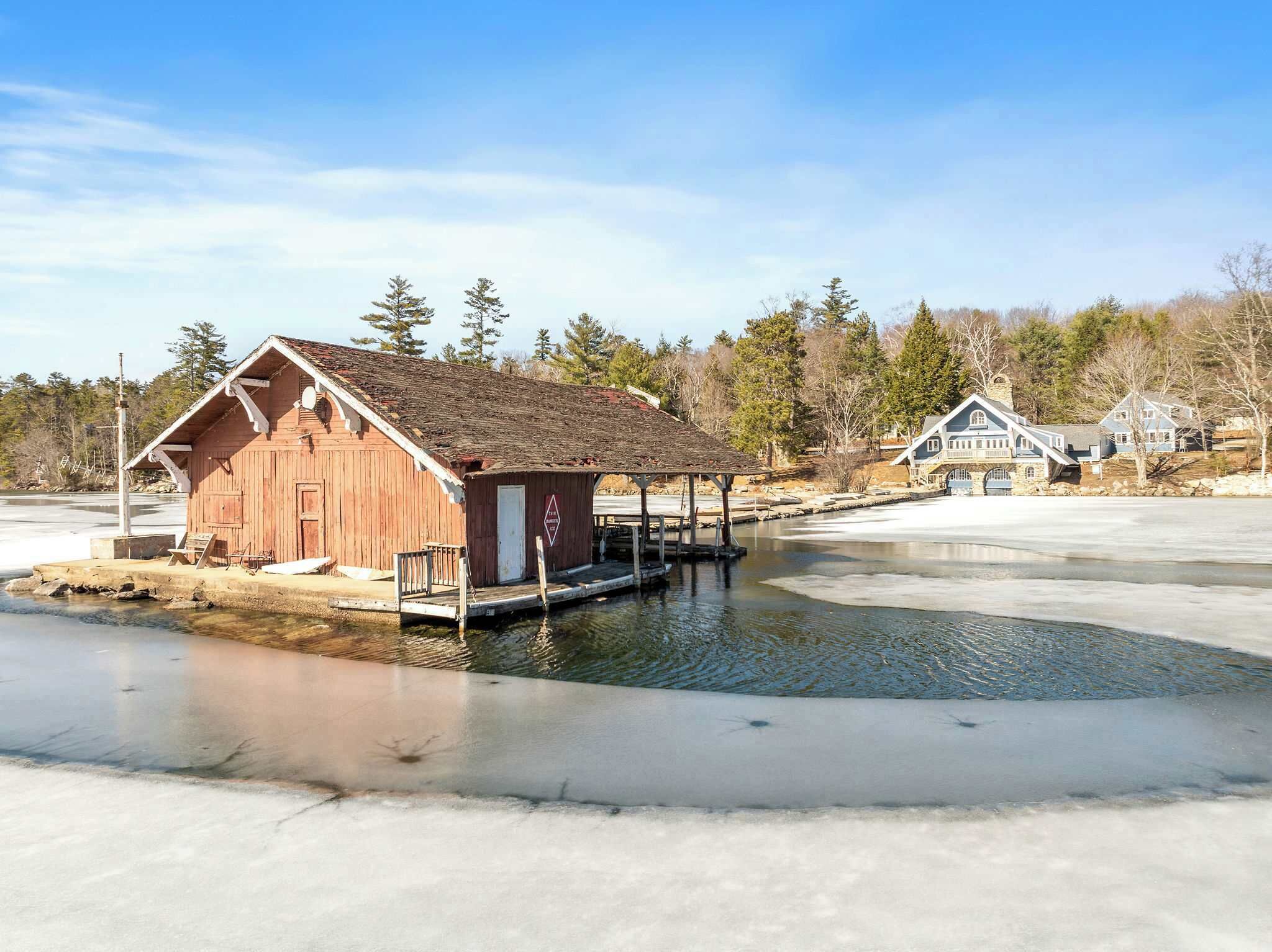 A wooden cabin with a dock extending into a partially frozen lake, surrounded by trees and houses.