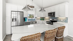 Une cuisine moderne avec des armoires blanches, des appareils en acier inoxydable, un grand îlot central, des chaises en osier et des luminaires suspendus.
