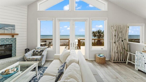 Un salon spacieux avec un haut plafond, de grandes fenêtres, une cheminée, un canapé et une vue sur la plage.