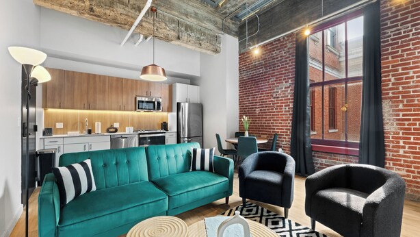 Una sala de estar moderna con un sofá azul verdoso, sillones negros y una mesa de centro de madera. Se observa una cocina con electrodomésticos de acero inoxidable al fondo.