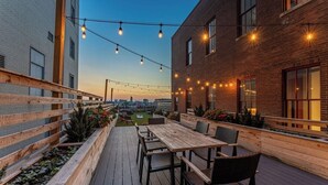 Un patio al aire libre con una terraza de madera, una mesa con sillas, luces colgantes y vistas de la ciudad.