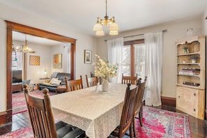 A dining room with a large table, chairs, a chandelier, a bookshelf, and a window with curtains.