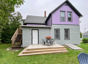 Ein zweistöckiges Haus mit grau-violettem Anstrich, einer Haustür, einer kleinen Terrasse mit Tisch und Stühlen und einer Holztreppe, die zu einer Veranda führt.